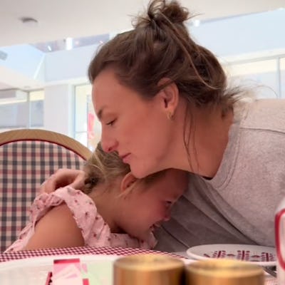 A woman gently embraces a young girl, both showing emotion, while seated at a table adorned with plates and condiments in a bright, airy setting.