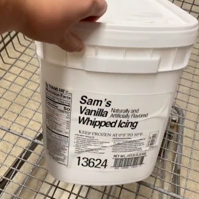 A person's hand holds a large white container of Sam's Vanilla Whipped Icing, labeled with nutritional information, in a shopping cart.