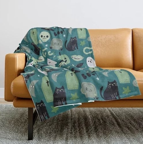 A cozy, patterned blanket featuring black cats, tombstones, and playful Halloween motifs draped over...