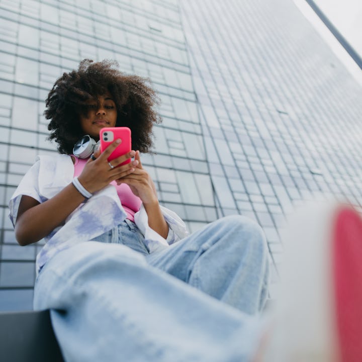 A young person with curly hair sits casually, using a pink phone. They wear a white shirt and baggy ...