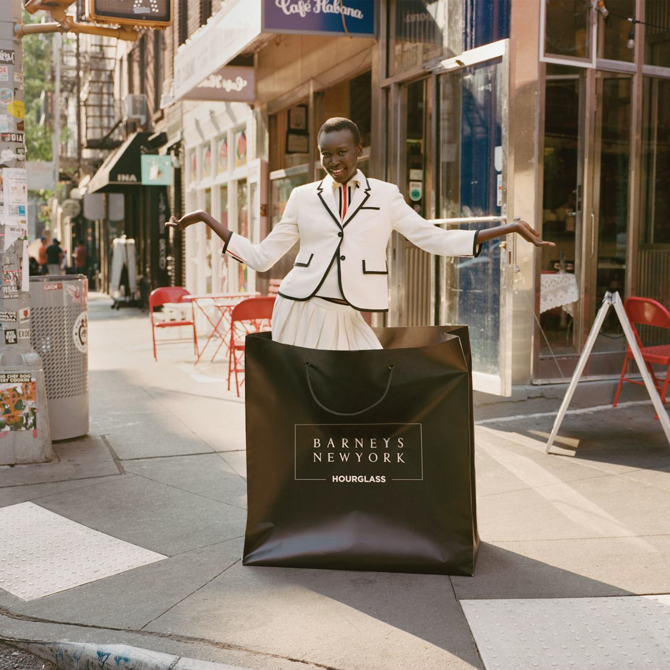 A fashionable individual stands on a city street, wearing a tailored white blazer and skirt, holding...