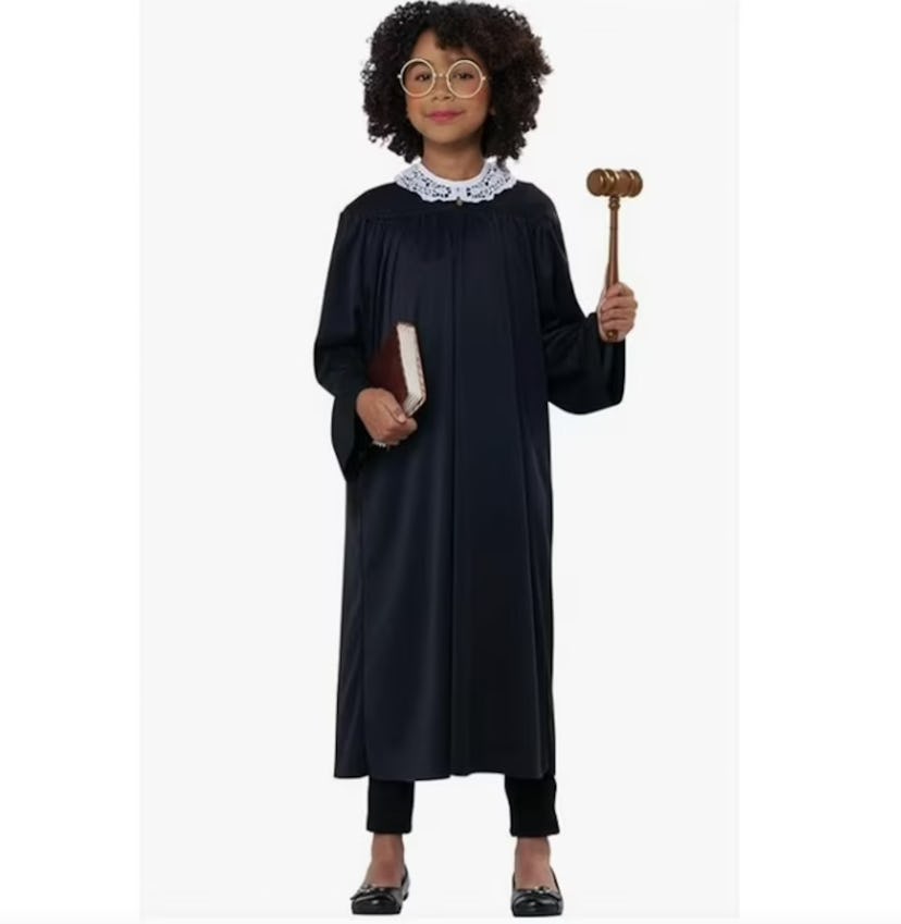 A young girl with curly hair wears a black robe and large glasses, holding a gavel in one hand and a...