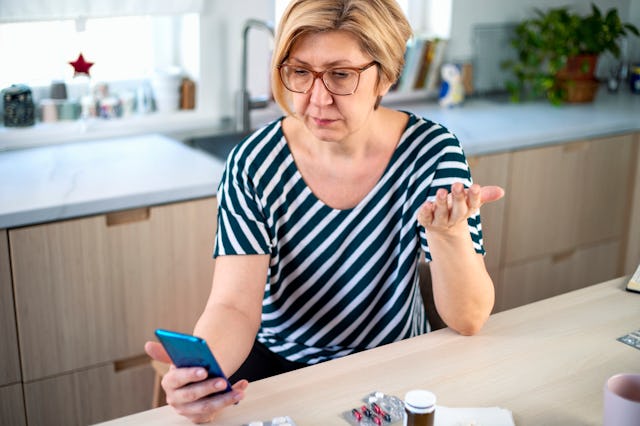 A middle-aged woman, wearing a striped shirt and glasses, sits at a kitchen table, looking at her ph...