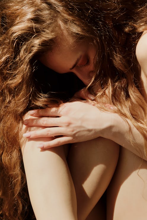 A woman with long, wavy hair rests her head on her knees, her arms wrapped around them in a contempl...