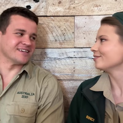 Chandler Powell and Bindi Irwin in zoo uniforms are sitting close together, smiling and looking at each other against a rustic wooden backdrop, conveying a friendly atmosphere.
