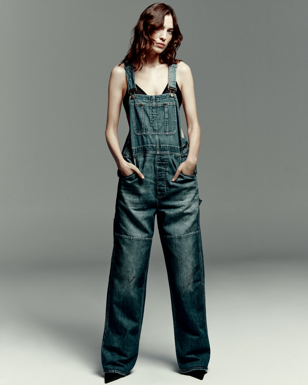 A young woman stands confidently in a pair of oversized denim overalls, hands in pockets, against a ...