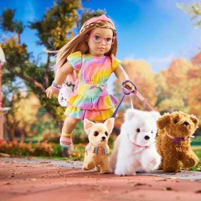 A colorful doll with braided hair, wearing glasses and a rainbow dress, walks two plush dogs on a leashed stroll through a sunny park.