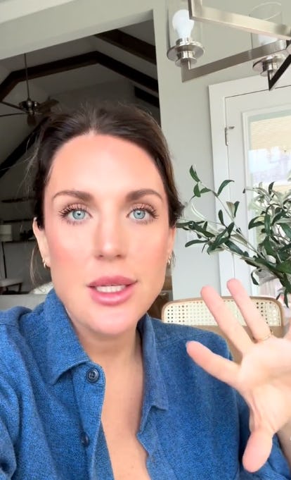 A woman with long brown hair and blue eyes is speaking directly to the camera. She wears a blue button-up shirt and gestures with her hand. A plant is visible in the background.