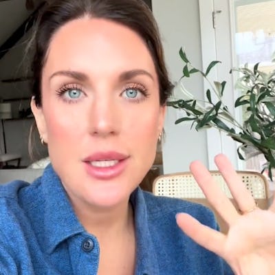 A woman with long brown hair and blue eyes is speaking directly to the camera. She wears a blue button-up shirt and gestures with her hand. A plant is visible in the background.