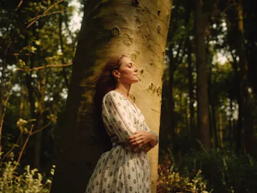 Kate Middleton in a flowing white dress leans against a large tree in a sunlit forest, eyes closed, ...