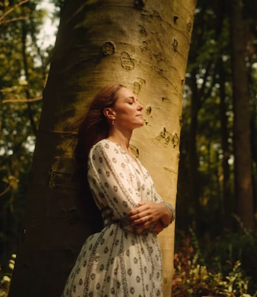 Kate Middleton in a flowing white dress leans against a large tree in a sunlit forest, eyes closed, ...