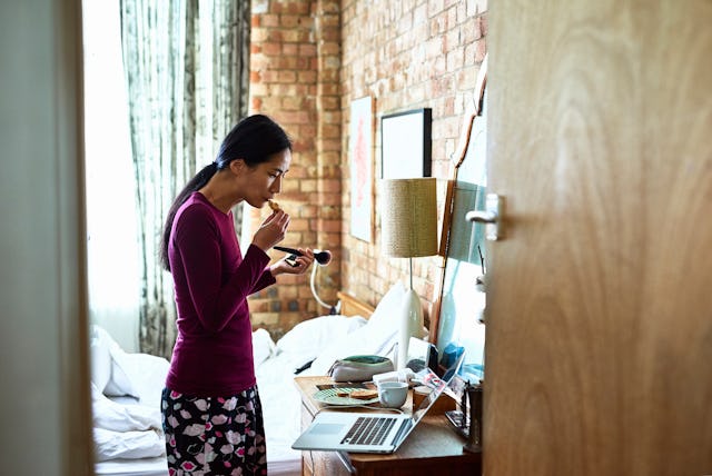 A woman stands in a cozy bedroom while applying makeup. The room features a brick wall, a messy bed,...
