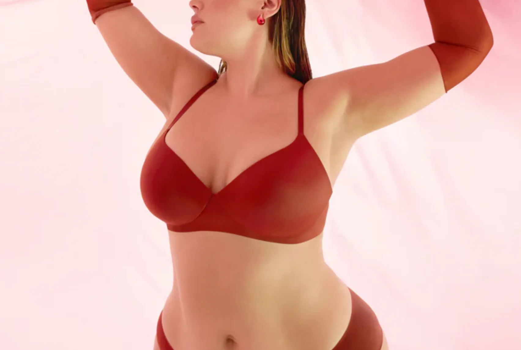 Model poses gracefully in a red bra and matching undies against a soft pink backdrop. 
