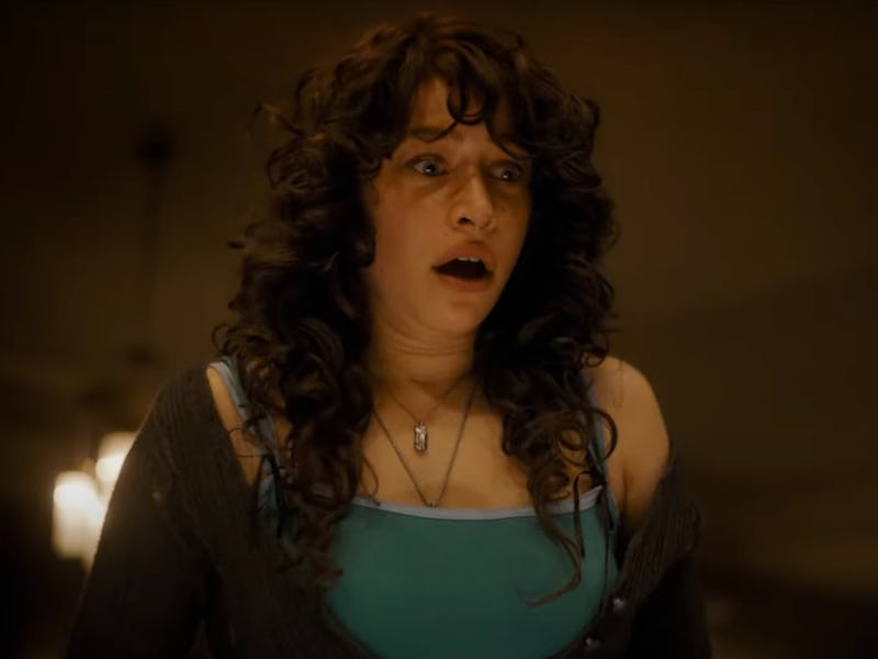A woman with curly hair looks shocked and distressed, wearing a blue top and a dark cardigan, standi...