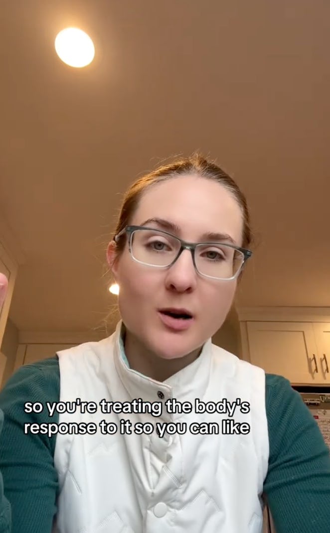 A person with glasses and light brown hair speaks intently, gesturing slightly. They wear a white vest over a green shirt, with a kitchen backdrop.
