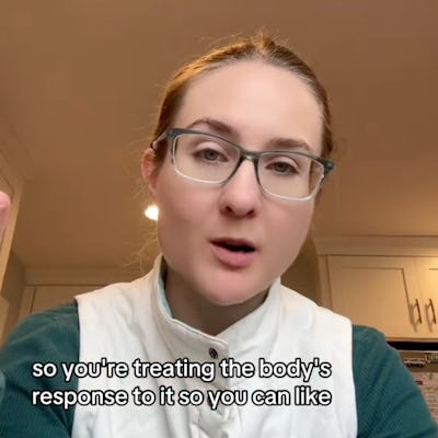 A person with glasses and light brown hair speaks intently, gesturing slightly. They wear a white vest over a green shirt, with a kitchen backdrop.