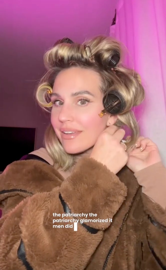 A woman with curly hair is smiling while wearing hair rollers, dressed in a cozy brown jacket, against a softly lit background with pink tones.