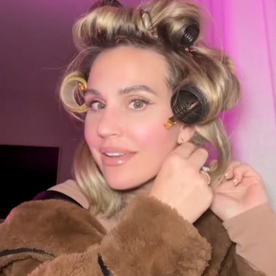 A woman with curly hair is smiling while wearing hair rollers, dressed in a cozy brown jacket, against a softly lit background with pink tones.