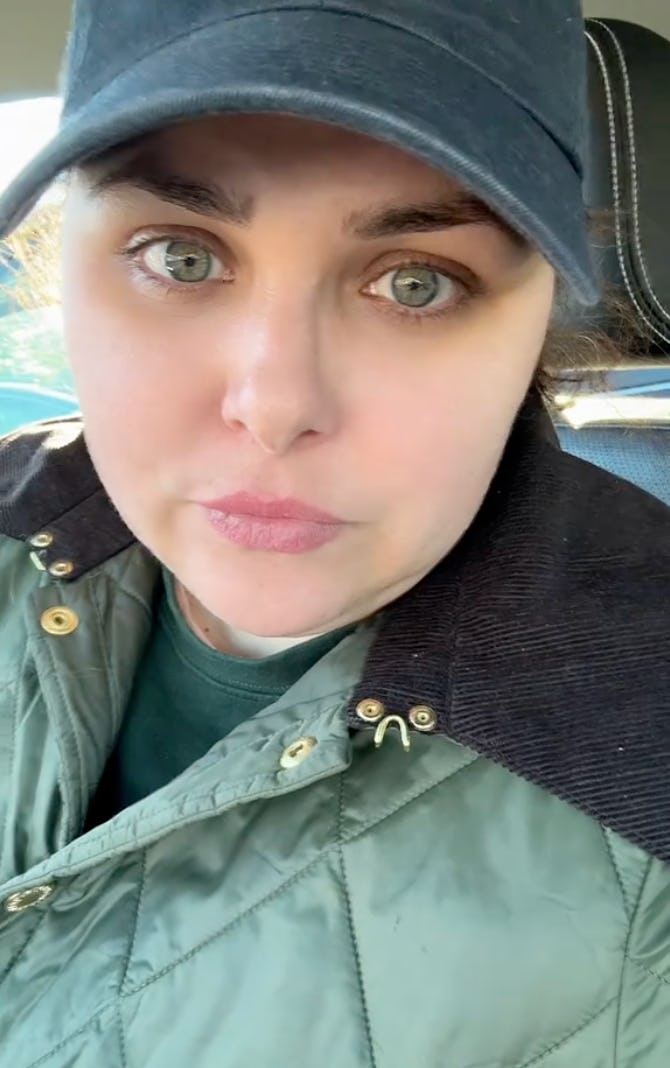 A close-up of a woman with green eyes, wearing a cap and a quilted jacket, looking directly at the camera from inside a car.
