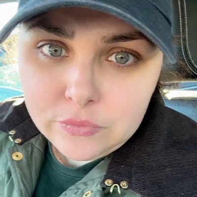 A close-up of a woman with green eyes, wearing a cap and a quilted jacket, looking directly at the camera from inside a car.