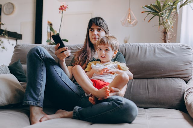 A woman sits on a couch, holding a smartphone while a young boy sits on her lap. They are both engag...