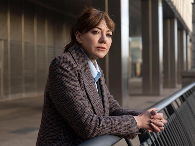 A woman in a patterned blazer leans against a railing, gazing thoughtfully. The modern architecture ...