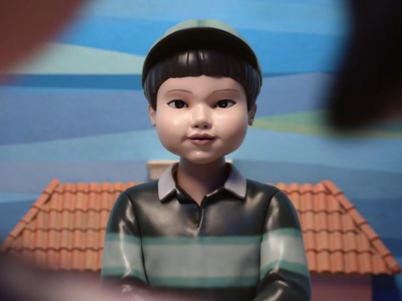 A close-up of a stylized doll wearing a striped shirt, seated against a colorful backdrop with a hou...