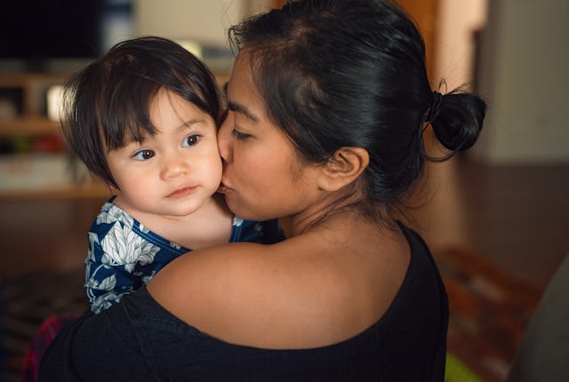 A woman gently kisses a toddler she holds in her arms. The scene is warm, capturing a moment of affe...