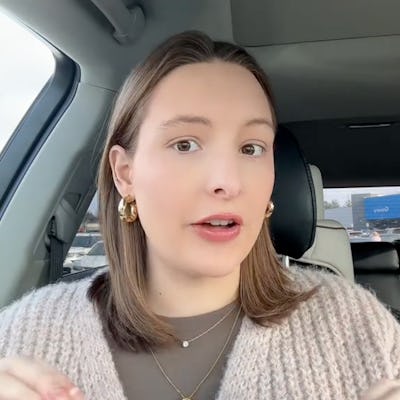 A woman with shoulder-length hair sits in a car, wearing a cozy sweater and gold hoop earrings, speaking expressively with a thoughtful expression.