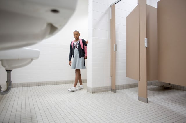 A young girl with a backpack stands in a school bathroom, gazing toward the stalls, with white tiles...