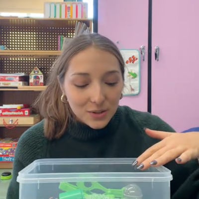 A young woman with brown hair sits in a classroom, engaging with a transparent container filled with green material and small toys.