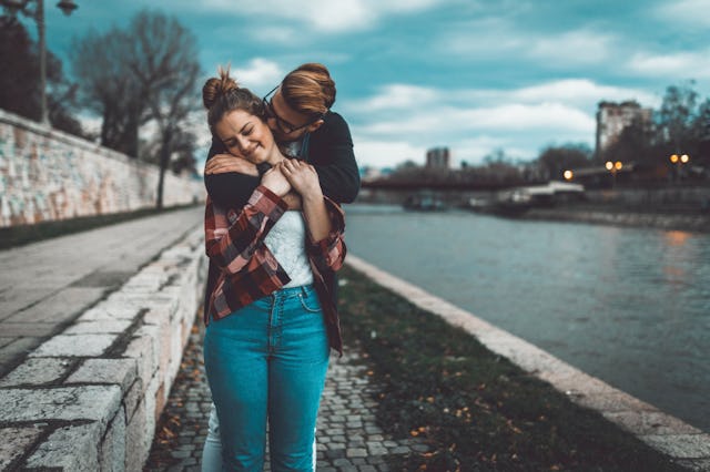 Two women embrace on a riverside path, one playfully hugging the other from behind. They smile as th...
