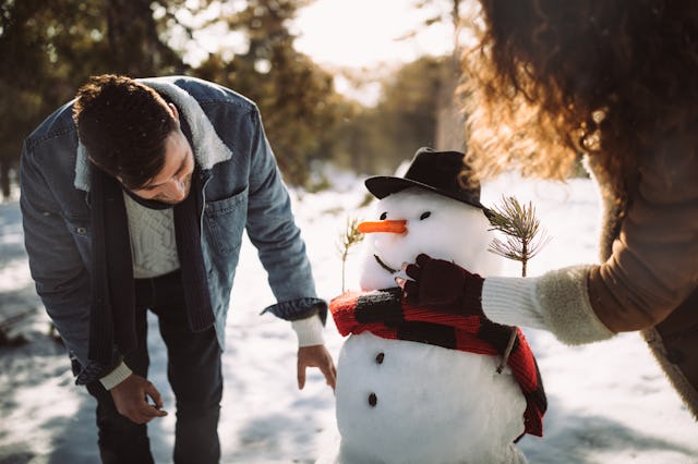Two people are decorating a snowman in a snowy landscape. One person adjusts the snowman's face whil...