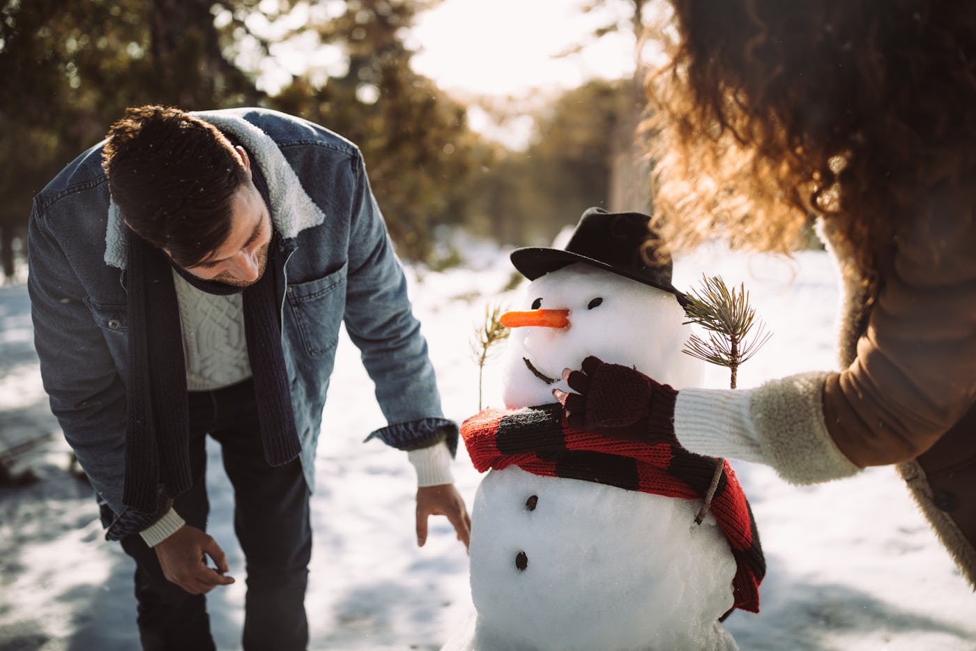Two people are decorating a snowman in a snowy landscape. One person adjusts the snowman's face while the other adds a hat and scarf.