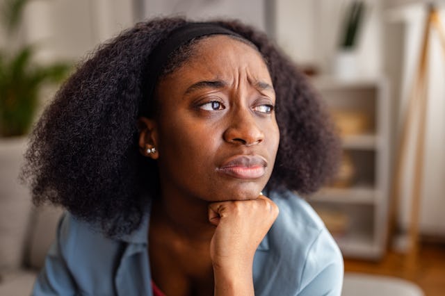 A woman with natural, curly hair looks pensive, resting her chin on her hand. She appears deep in th...