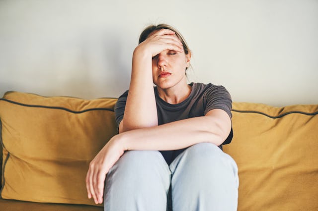 A young woman sits on a yellow couch, her head resting on her hand in a gesture of distress, conveyi...