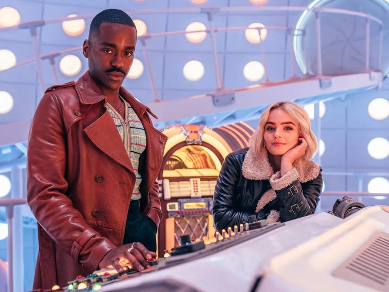 Two characters stand in a colorful control room filled with circular lights. One man in a brown coat...