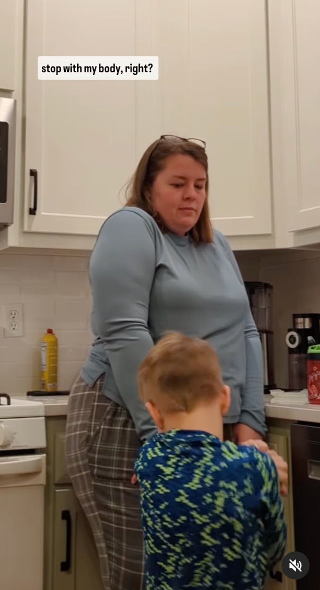 A woman stands in a kitchen wearing a blue top and plaid pants, looking contemplative, while a young boy, dressed in a colorful sweater, interacts with her.