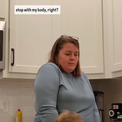 A woman stands in a kitchen wearing a blue top and plaid pants, looking contemplative, while a young boy, dressed in a colorful sweater, interacts with her.