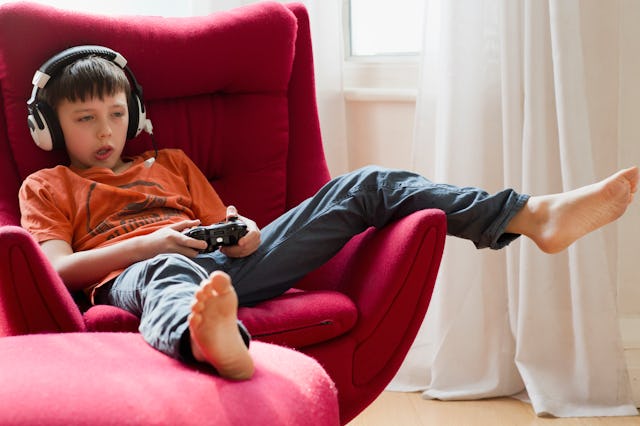 A boy sits comfortably in a bright red chair, wearing headphones and playing a video game with a con...