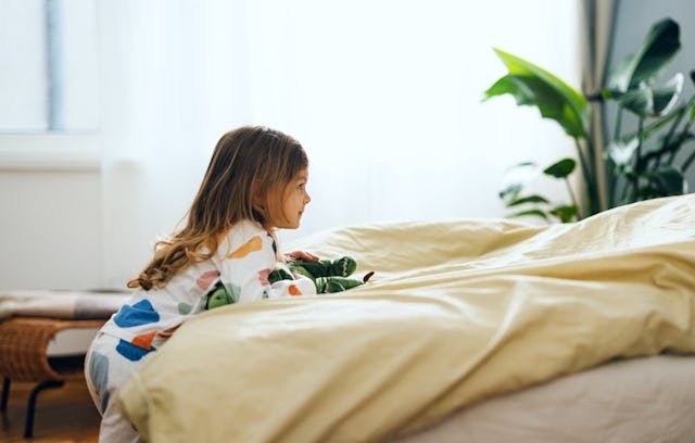 A young girl in colorful pajamas leans over a bed, peering intently at something hidden beneath yell...