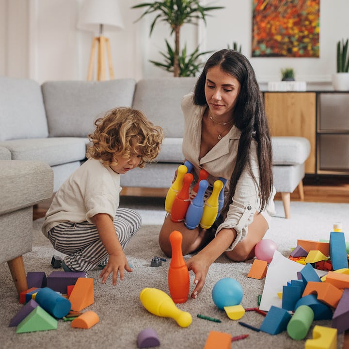 A woman sits on a rug, holding colorful bowling pins, while a young boy kneels beside her, surrounde...