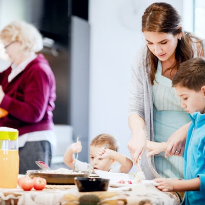A woman assists two children with cooking at a kitchen table while another woman prepares food in the background. Fresh ingredients are visible.