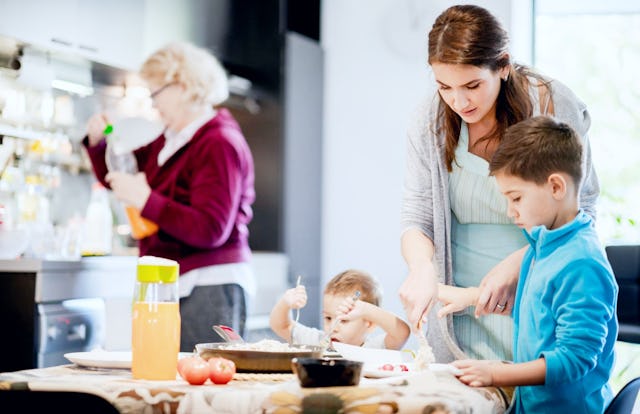 A woman assists two children with cooking at a kitchen table while another woman prepares food in th...
