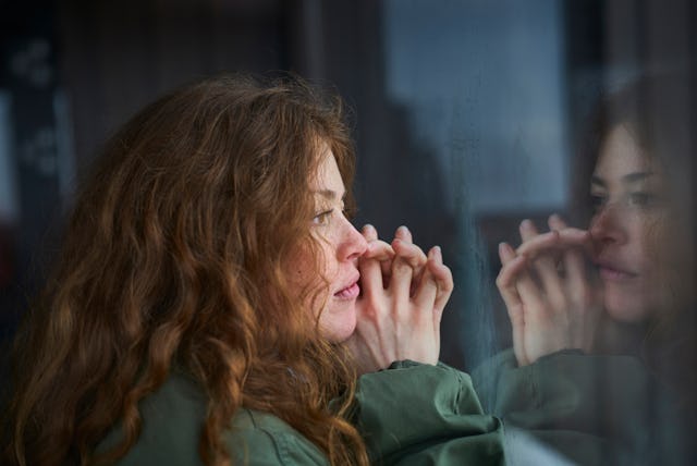 A woman with long, curly hair gazes thoughtfully out a window, her hands pressed against the glass, ...