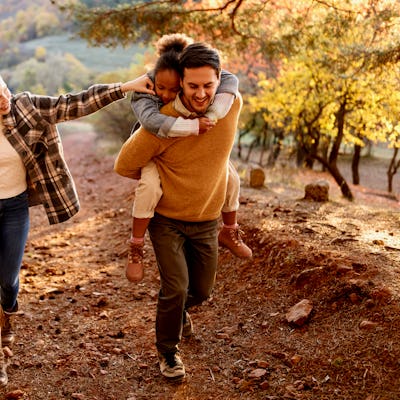 A woman gets to know her stepdaughter on a sunny autumn day outdoors, with a girl on her father's back and the woman playfully holding his arm while walking on a dirt path.