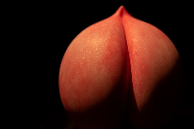 A close-up image of a smooth peach, resembling a pair of rounded forms, illuminated against a dark b...