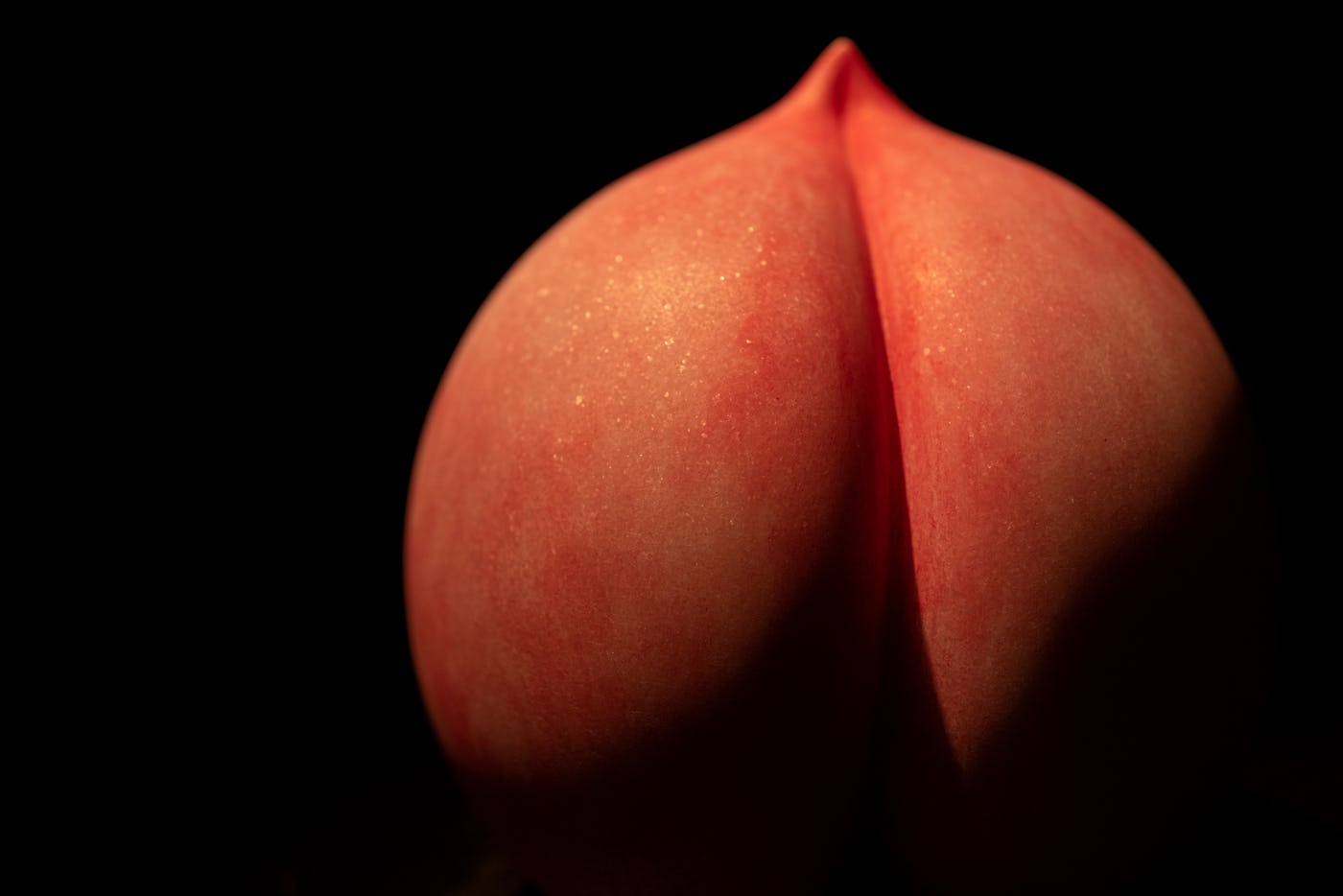 A close-up image of a smooth peach, resembling a pair of rounded forms, illuminated against a dark background.