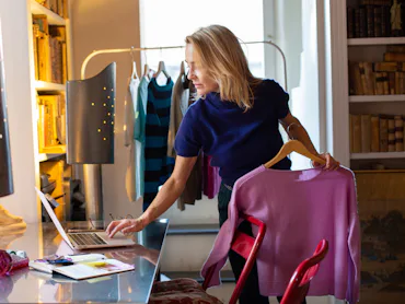 A woman in a navy top stands at a desk, checking a laptop while holding a pink sweater. Colorful clo...