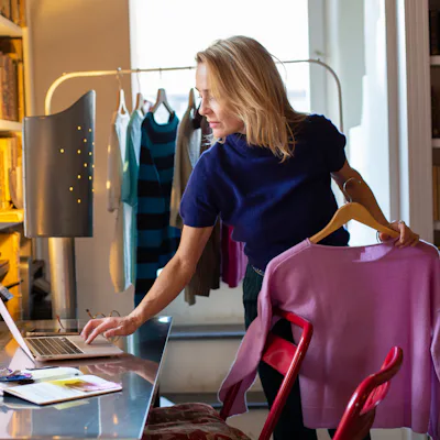 A woman in a navy top stands at a desk, checking a laptop while holding a pink sweater. Colorful clothing hangs in the background, creating a vibrant workspace.